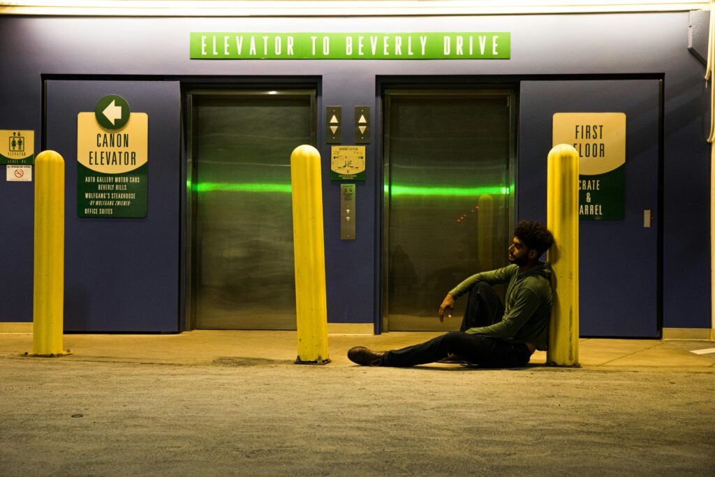 A man sitting by an elevator entrance under neon lights at night.