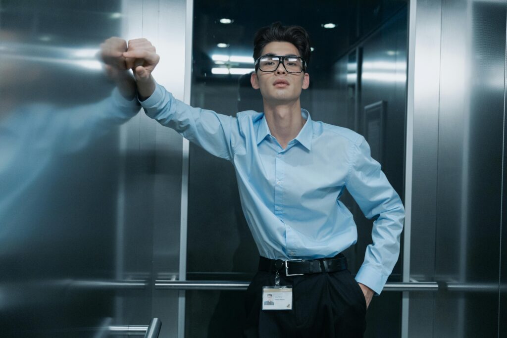 Portrait of an Asian man in blue long sleeves with eyeglasses, leaning in an elevator.